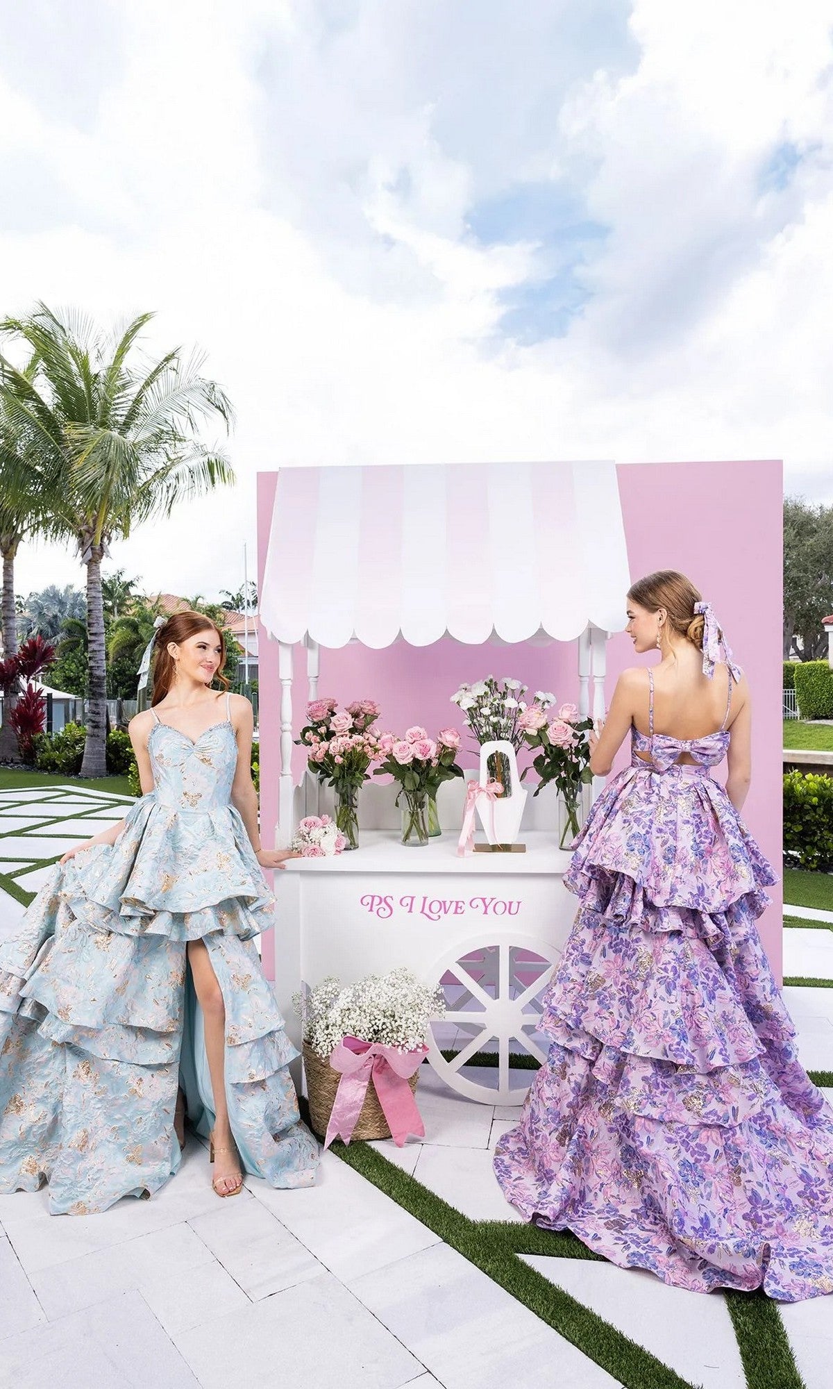 Two women in tiered floral dresses, including the Long Formal Dress: Portia and Scarlett XO181 with its high-low design, stand by a pink and white flower cart labeled